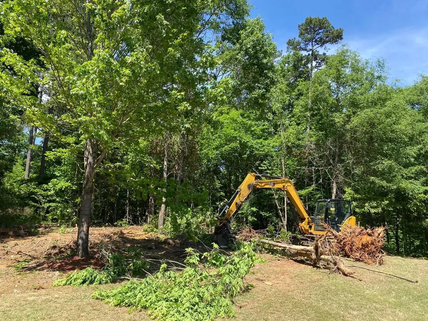Excavator pulling small trees and brush at the edge of a cleared yard, land clearing