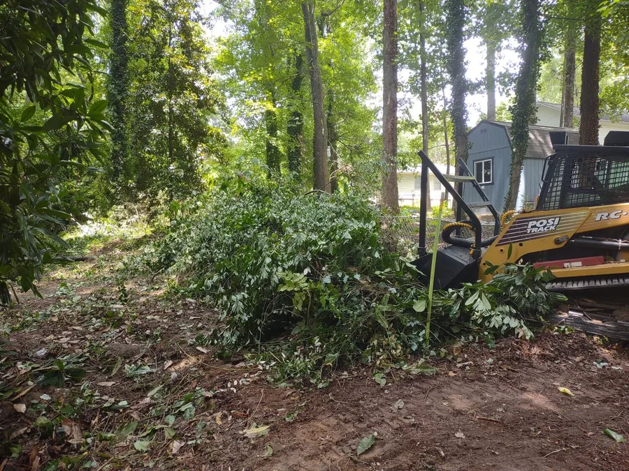 Tracked skid loader with a mulching head beside a pile of cut brush near a backyard shed, forestry mulching