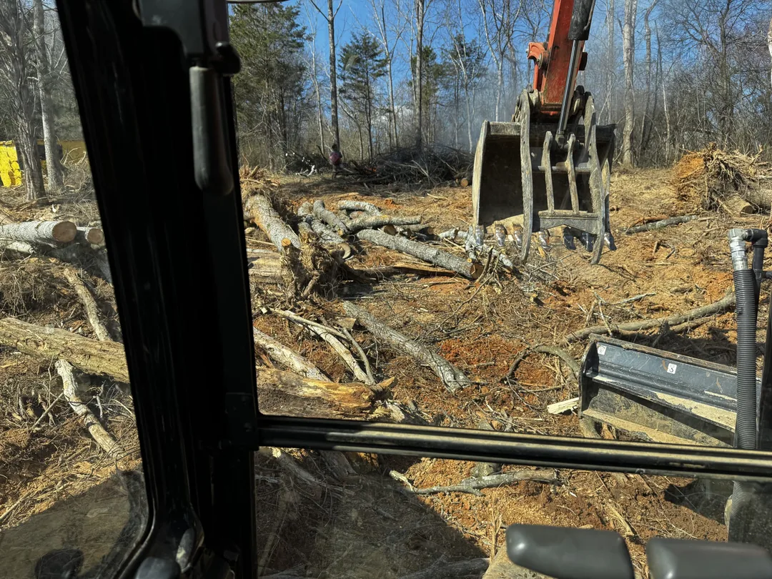 Excavator with a thumb clearing felled trees and brush on a wooded lot, seen from the cab, lot clearing