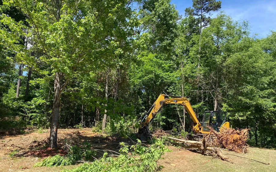 Excavator pulling small trees and brush at the edge of a cleared yard, lot clearing