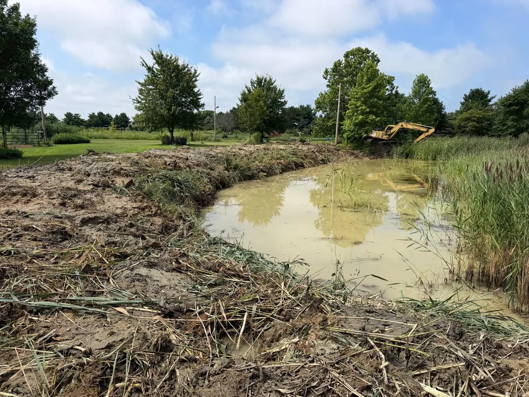 Freshly cleaned out farm pond with dug muck along the banks and an excavator on the far bank, pond cleaning