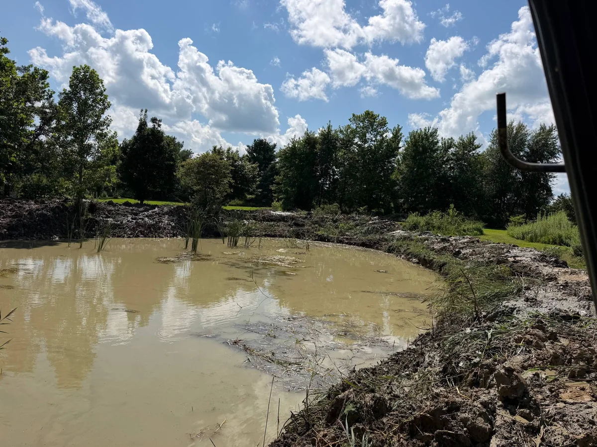 Cleaned out pond seen from the excavator cab with fresh muck banks along the shoreline, pond cleaning