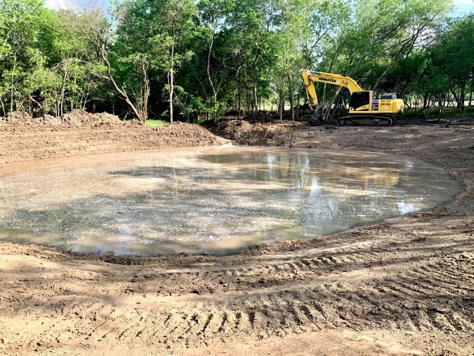 Excavator beside a newly dug pond basin with fresh dirt banks, pond construction