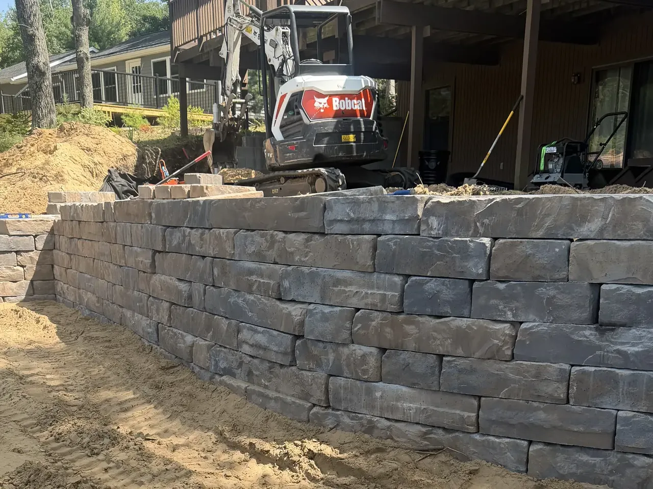 Block retaining wall under construction with a mini excavator behind it, retaining walls