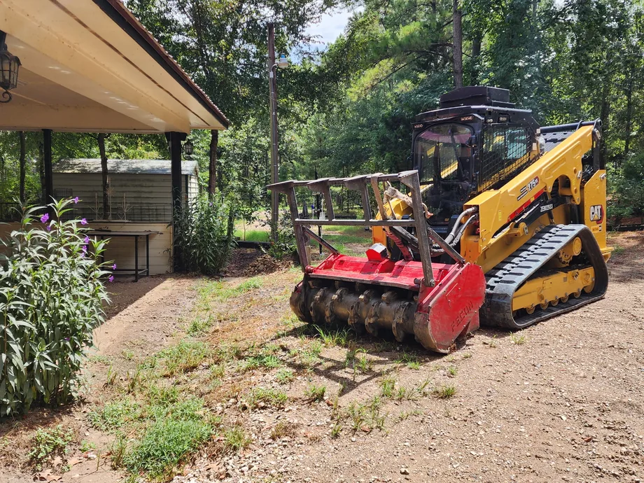 Tracked skid loader with a mulching head on cleared ground under shade trees, site preparation