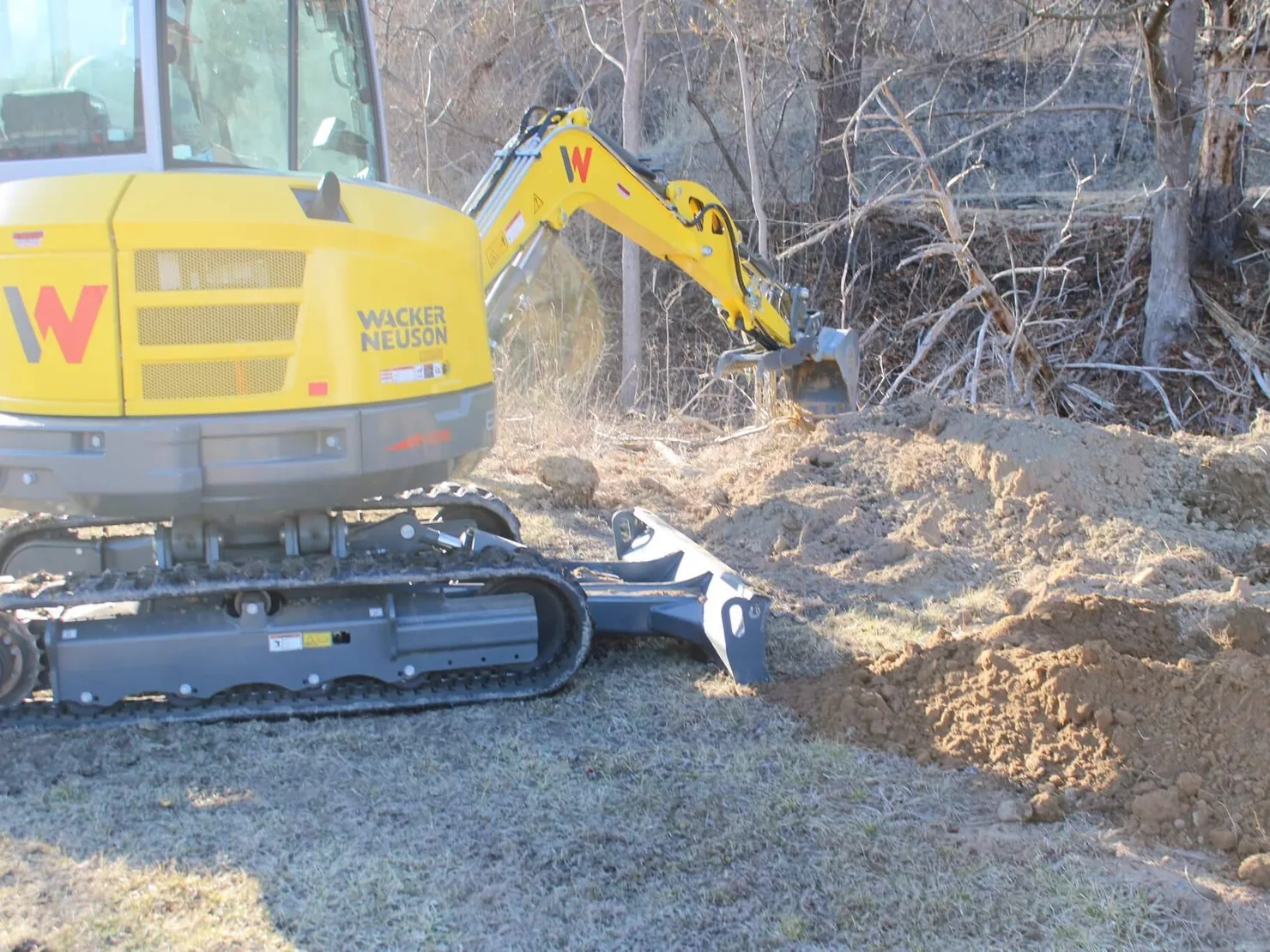 Mini excavator cutting into a dirt bank and leveling soil at the edge of a brushy lot, site preparation